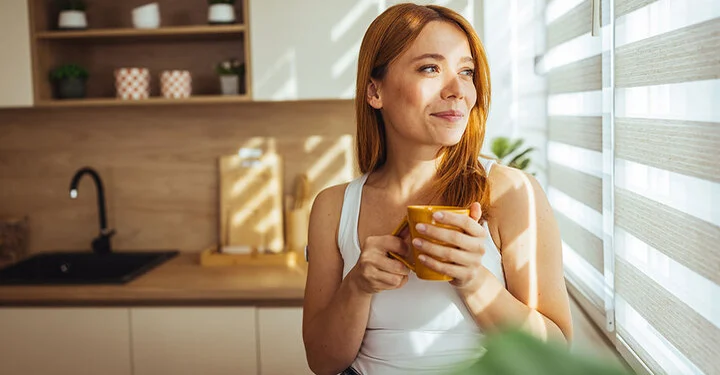 Frau vor einer Innenjalousie in einer gut beschatteten Küche mit modernem Sonnenschutz, hält eine Tasse und schaut lächelnd hinaus.