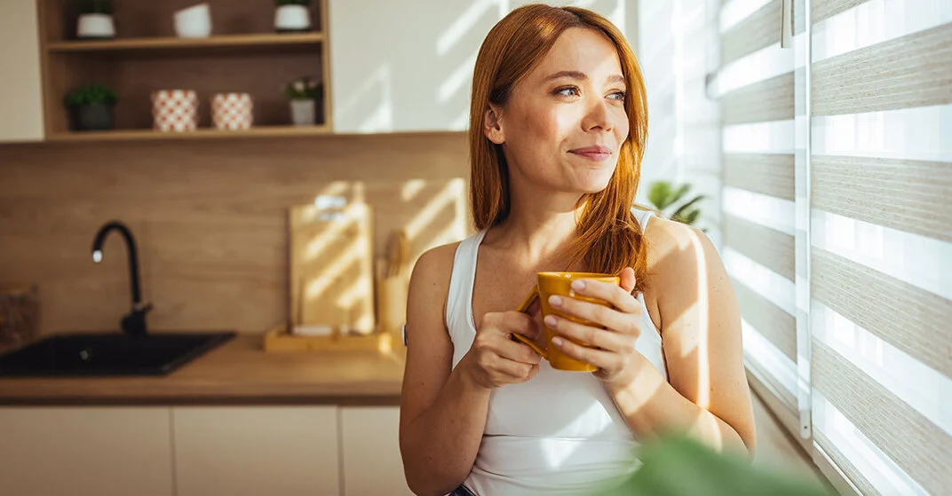 Frau vor einer Innenjalousie in einer gut beschatteten Küche mit modernem Sonnenschutz, hält eine Tasse und schaut lächelnd hinaus.