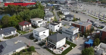 Mehrere moderne Einfamilienhäuser in verschiedenen Designs stehen in einem dicht bebauten Wohngebiet. Im Hintergrund befindet sich ein großes Einkaufszentrum mit vielen geparkten Autos und grünen Bäumen.