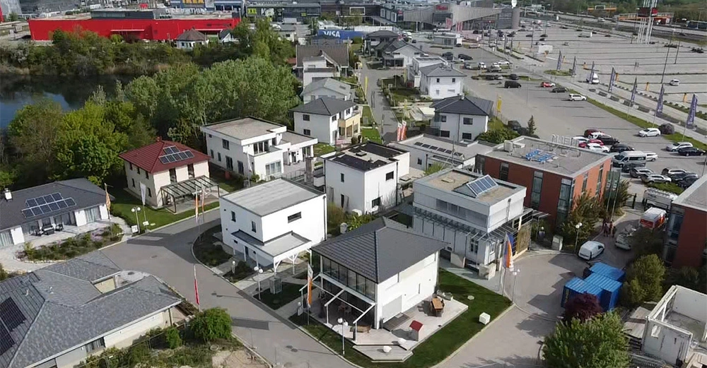 Mehrere moderne Einfamilienhäuser in verschiedenen Designs stehen in einem dicht bebauten Wohngebiet. Im Hintergrund befindet sich ein großes Einkaufszentrum mit vielen geparkten Autos und grünen Bäumen.