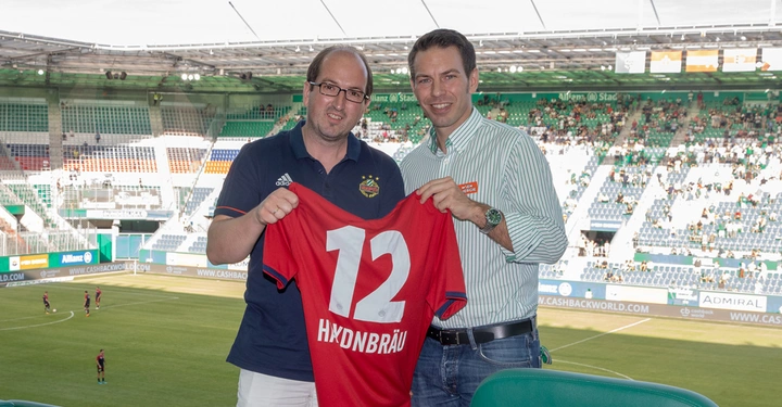 Zwei Männer halten zusammen ein rotes Trikot mit der Nummer 12 und dem Schriftzug HXDNBRÄU Stadion im Hintergrund zuschauende Menge und Fußballspieler auf dem Spielfeld.