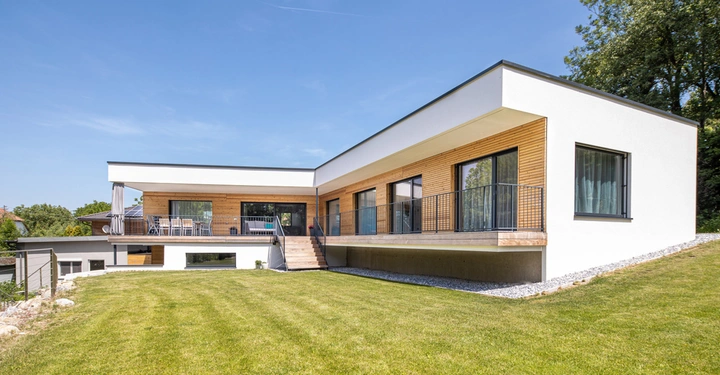 Ein modernes einstöckiges Haus steht auf einer Rasenfläche Treppe und Balkon im Vordergrund umgeben von Bäumen und klarem Himmel. Hauswände aus Holz- und weißem Putz.