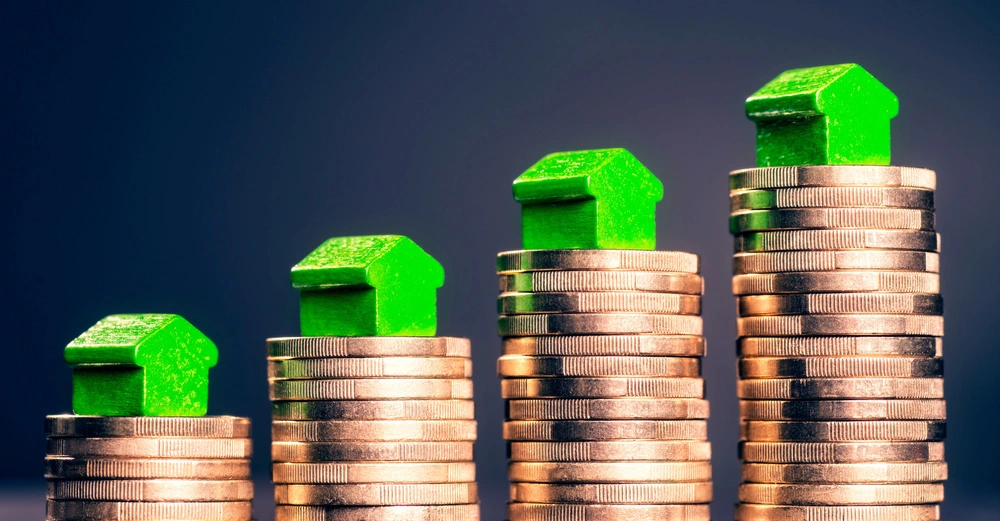 Grüne Spielzeughäuser stehen auf stapelweise Münzen in ansteigender Reihenfolge vor einem dunklen Hintergrund