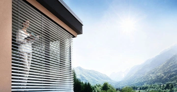 Frau steht vor halbgeöffnetem Raffstore und sieht entspannt auf die idyllische Berglandschaft vor ihrem Haus.