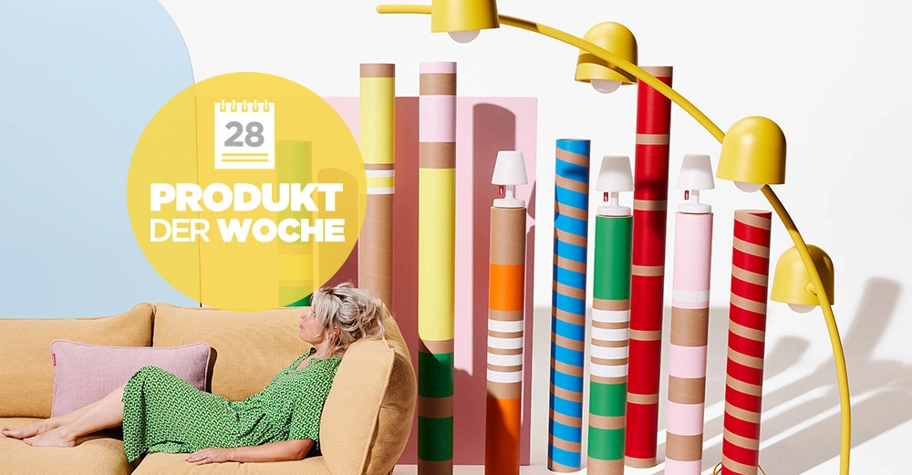 Frau sitzt auf gelbem Sofa, umgeben von bunten, gestreiften Zylindern und gelber Stehlampe. Links ist eine gelbe Kreisgrafik mit der Aufschrift: 28 PRODUKT DER WOCHE.