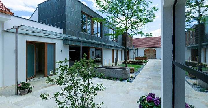 Galerie-Bild 7: Moderner Innenhof mit Pflanzenbeeten und Steinwegen zwischen Gebäuden mit großen Fenstern und verglasten Türen; Bäume und blauer Himmel im Hintergrund.