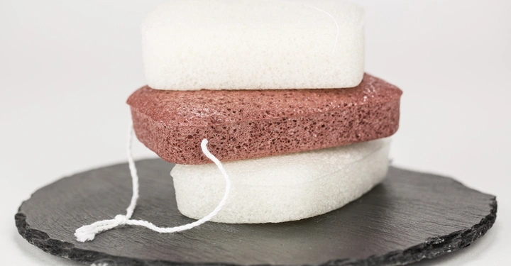 Galerie-Bild 5: Drei Badeschwämme sind aufeinander gestapelt auf einem runden Schieferplatte und haben weiße Schnüre, die lose herunterhängen. Der Hintergrund ist weiß und schlicht.