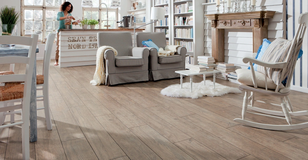 Eine Frau schneidet Gemüse in einer hellen modernen Wohnküche mit Holzboden zwei graue Sessel ein Bücherregal ein Kamin mit Kerzen und einem Schaukelstuhl SEAN TIKY NORD 172 ist an der Theke geschrieben