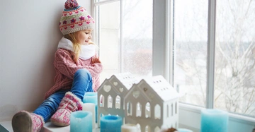 Ein kleines Mädchen mit Mütze und Schal sitzt lächelnd auf einer Fensterbank Jeans und Winterstiefel tragend. Davor stehen blaue Kerzen und weiße Dekorhäuser mit Schneeflockendesign draußen kahler Baum.