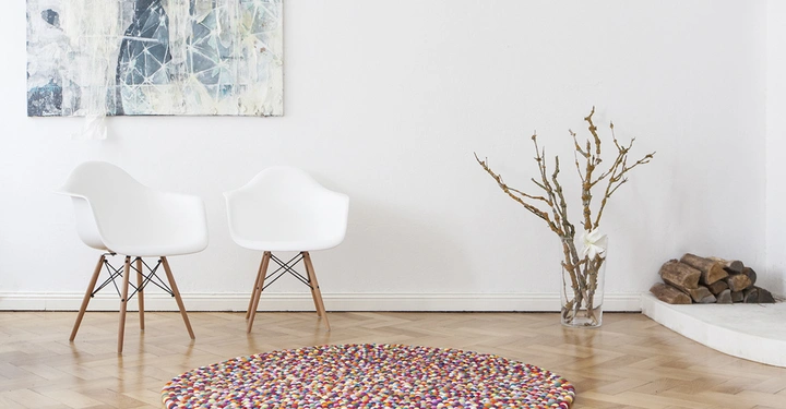 Galerie-Bild 5: Zwei weiße Stühle stehen nebeneinander auf einem Holzfußboden. Im Hintergrund hängt ein abstraktes Gemälde an der weißen Wand. Rechts steht ein Glasgefäß mit Zweigen. Ein bunter runder Teppich liegt auf dem Boden. Das Bild zeigt außerdem einen kleinen Stapel Brennholz in der Ecke.