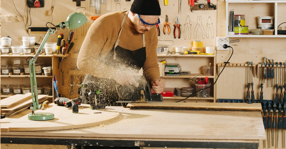 Person arbeitet mit einer elektrischen Säge in einer Holzwerkstatt Werkzeuge und Materialien sind an der Wand und auf Regalen organisiert Eine grüne Lampe und Bohrer befinden sich auf dem Tisch
