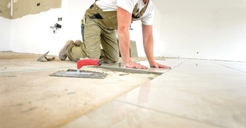 Handwerker in grau-grüner Latzhose kniet am Boden und drückt eine Bodenfliese in den Mörtel