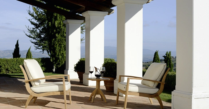 Zwei gepolsterte Stühle stehen auf einer Terrasse unter einem Holzdach vor weißen Säulen Ein kleiner runder Tisch dazwischen trägt zwei Weingläser und eine Weinflasche Im Hintergrund ist ein Garten zu sehen