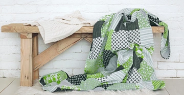 Holzbank mit weißem Strickpullover und grün-gemusterter Patchwork-Decke darauf liegend vor weißer Ziegelwand in einem hellen Raum auf einem weißen Teppich.