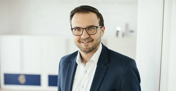 Mann lächelt leicht während er in die Kamera schaut trägt Anzug und Brille im gut beleuchteten Büro mit verschwommenem Hintergrund.