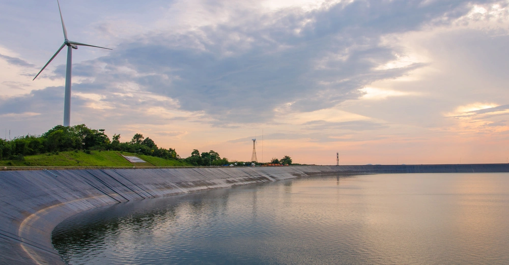 Windrad stehend auf grünem Hügel dreht sich langsam neben einem großen künstlichen Wasserreservoir unter bewölktem, teils sonnigem Himmel.