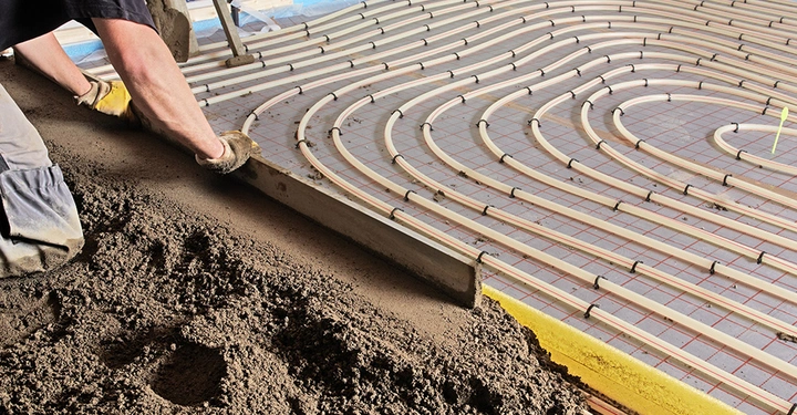 Ein Handwerker bringt den Heizestrich über den verlegten Rohren der Fußbodenheizung auf. Der Estrich dient als Wärmespeicher und sorgt für eine gleichmäßige und angenehme Strahlungswärme.