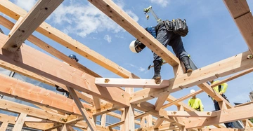 Arbeiter bauen auf einem Holzgerüst und verwenden Hämmer bei sonnigem Wetter und blauem Himmel im Hintergrund. Weitere Arbeiter tragen Schutzhelme und fluoreszierende Hemden.