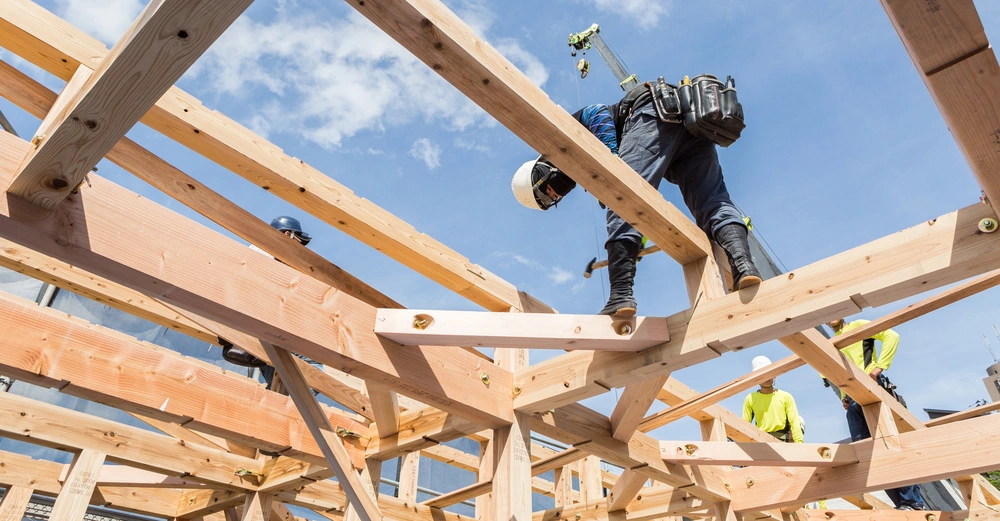 Arbeiter bauen auf einem Holzgerüst und verwenden Hämmer bei sonnigem Wetter und blauem Himmel im Hintergrund. Weitere Arbeiter tragen Schutzhelme und fluoreszierende Hemden.