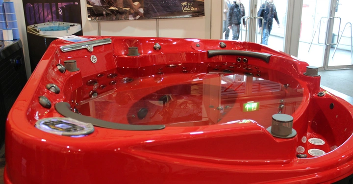 Galerie-Bild 23: Rotes Whirlpool steht still auf Ausstellung Messestand links Poster mit Menschen Link zur Marke Coast Spas Taking it to the Edge Quality. Im Hintergrund Messeeingang mit Personen.