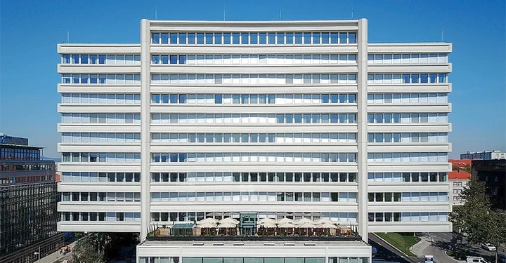 Ein großes modernes Bürogebäude steht in einer städtischen Umgebung während des Tages Es hat viele Fenster und eine Dachterrasse mit Sonnenschirmen und Tischen unten sind Bäume und Straßen erkennbar