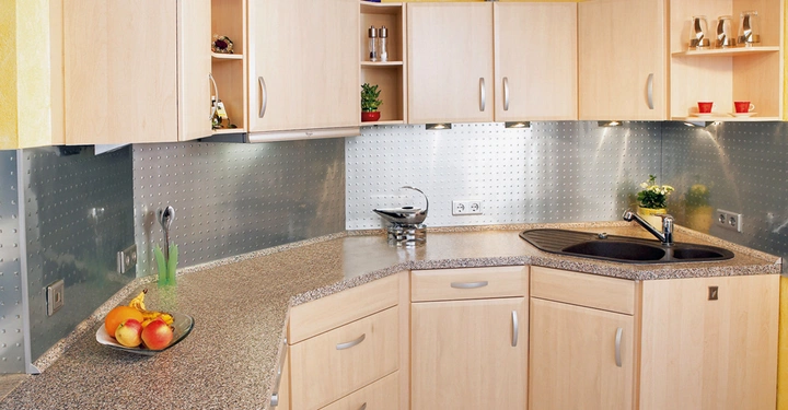 Eine moderne Küche verfügt über helle Holzschränke, eine graue Arbeitsplatte und eine Edelstahlrückwand. Auf der Arbeitsplatte befinden sich Obst, Kräuter und Küchenutensilien. Der Raum ist gut beleuchtet.