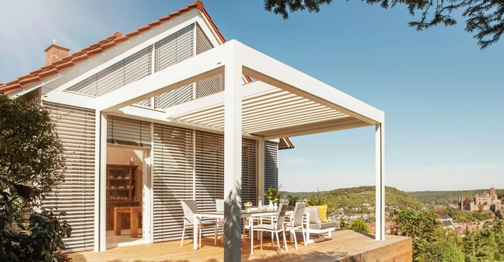 Weiße Pergola auf Holzterrasse neben modernem Haus mit Fensterläden und Blick auf hügelige Landschaft mit einem Schloss im Hintergrund bei sonnigem Wetter.