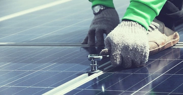 Handschuhtragende Hände befestigen eine Schraube auf einer Solarpanel-Oberfläche mit einem Schraubenschlüssel im Freien.