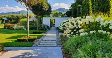 Gartenweg aus Steinplatten führt durch gepflegten Garten mit üppigen Blumen und Sträuchern Bäume werfen Schatten Berge und blauer Himmel im Hintergrund einladender friedlicher Ort