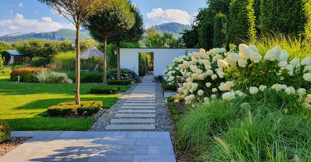 Gartenweg aus Steinplatten führt durch gepflegten Garten mit üppigen Blumen und Sträuchern Bäume werfen Schatten Berge und blauer Himmel im Hintergrund einladender friedlicher Ort