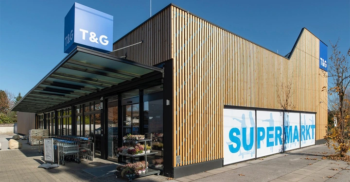 Galerie-Bild 1: Gebäude zeigt die Fassade eines Supermarkts aus Holz mit großen Fenstern und Glasvordach T&G-Logo auf blauem Schild Einkaufwagen draußen Pflanzen daneben Schriftzug SUPERMARKT auf der Wand.