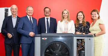 3 Männer in Anzug und 3 Frauen in Businesskleidung stehen lächelnd nebeneinander auf einer knallroten Bühne mit rotem Hintergrund. Vor ihnen eine Wärmepumpe von WOLF Österreich.