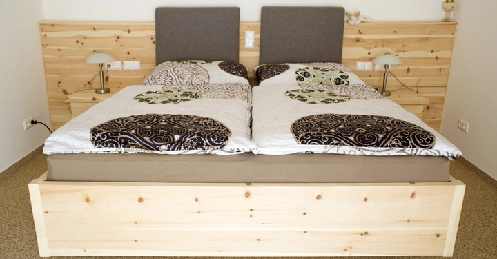 Galerie-Bild 8: Doppelbett mit gemusterten Bettdecken befindet sich in einem Schlafzimmer mit Holzwänden und Teppichboden. Zwei Nachttische mit Lampen und Steckdosen befinden sich neben dem Bett.