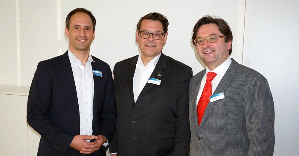Drei Männer stehen nebeneinander lächeln in die Kamera und tragen Namensschilder in einem hell erleuchteten Raum mit weißen Wänden. Die Namensschilder sind leider nicht lesbar.