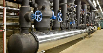 Große industrielle Rohre ausgestattet mit blau und schwarz lackierten Ventilen verlaufen durch einen Maschinenraum. Die Umgebung ist sauber und gut beleuchtet mit gut sichtbaren technischen Installationen.