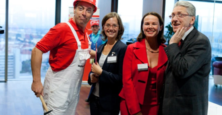 Galerie-Bild 43: Ein Mann in Arbeiterkleidung und Schutzhelm lächelt und zeigt auf drei geschäftlich gekleidete Personen die neben ihm stehen. Sie befinden sich in einem modernen Bürogebäude mit großen Fenstern und Stadtansicht.