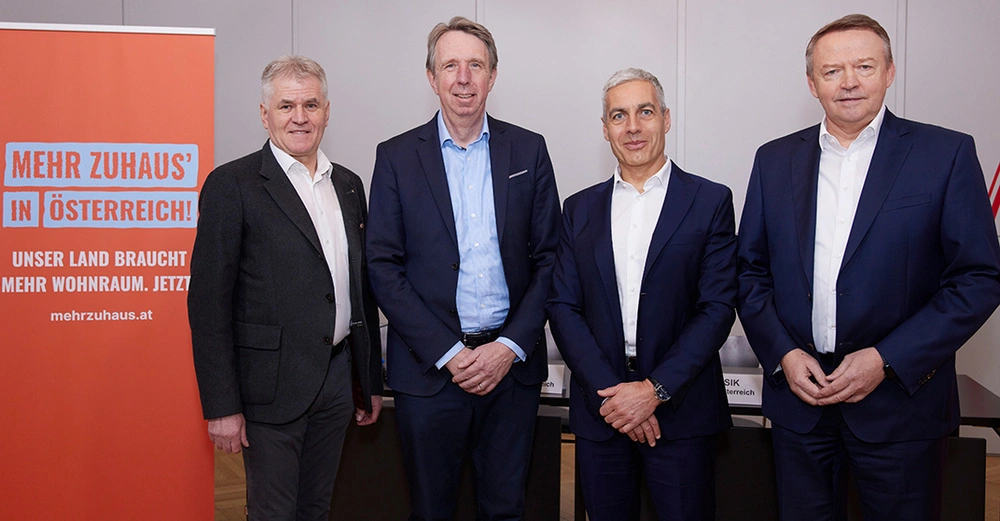 vier Männer in schwarzen Anzügen stehen lächelnd auf Bühne. Links im Bild ein großer Plakatständer mit der Aufschrift "Mehr Zuhaus in Österreich".