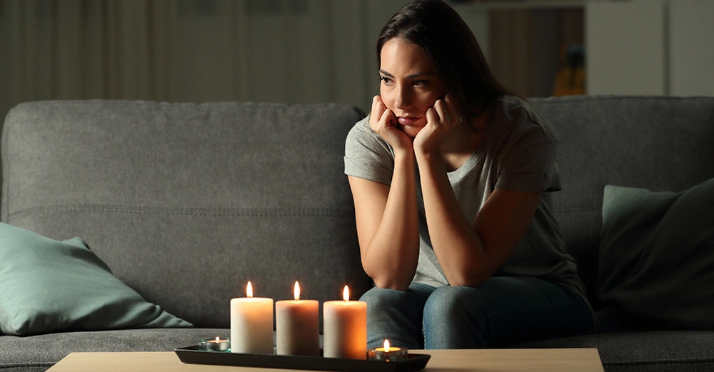 Frau sitzt nachdenklich auf einem grauen Sofa mit blaugrünen Kissen und schaut auf brennende Kerzen auf einem Couchtisch in einem gedämpft beleuchteten Raum