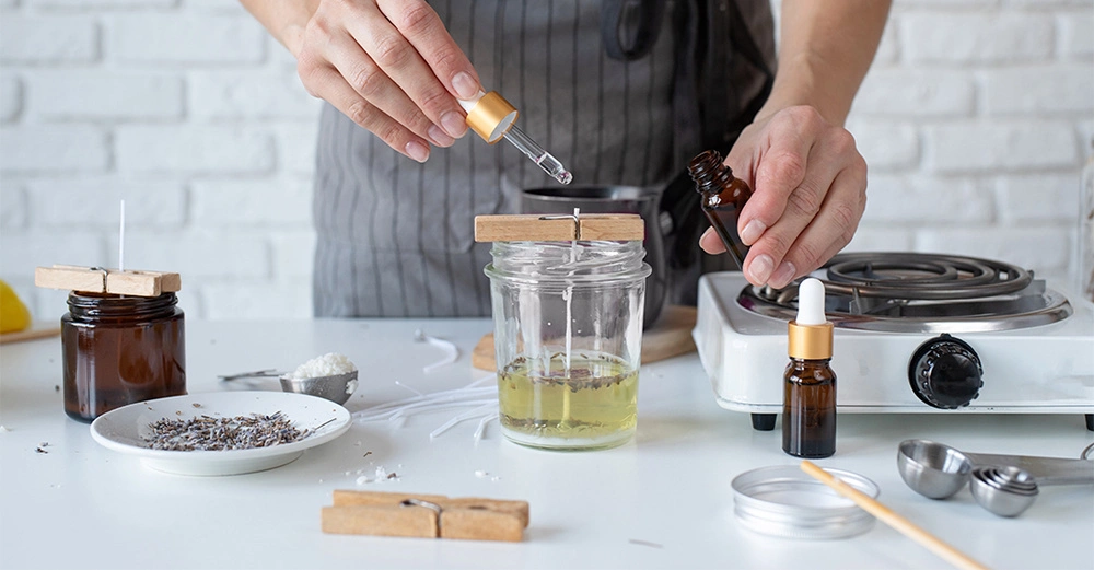 Frau tropft ätherisches Öl in eine Wachsschale, um Duftkerzen selbst herzustellen. Weiteres Zubehör und Herdplatte befinden sich auch auf dem Tisch.