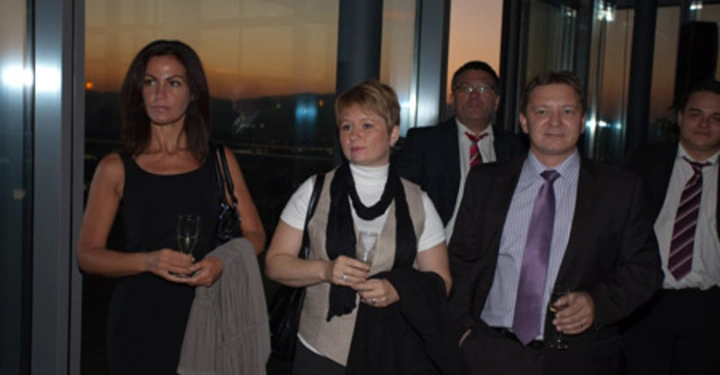 Galerie-Bild 67: Mehrere Personen stehen zusammen und halten Gläser in den Händen. Sie befinden sich in einem Raum mit großen Fenstern und Blick auf einen Sonnenuntergang.