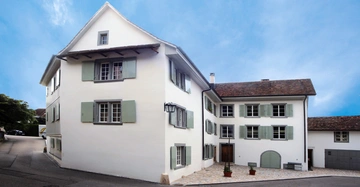 Zweistöckiges weißes Gebäude mit grünen Fensterläden an einer Straßenkreuzung bei klarem Himmel mehrere Fenster mit teilweise geöffneten Fensterläden und zwei Pflanzkübel vor dem Eingang.