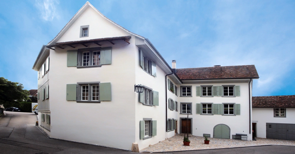 Zweistöckiges weißes Gebäude mit grünen Fensterläden an einer Straßenkreuzung bei klarem Himmel mehrere Fenster mit teilweise geöffneten Fensterläden und zwei Pflanzkübel vor dem Eingang.