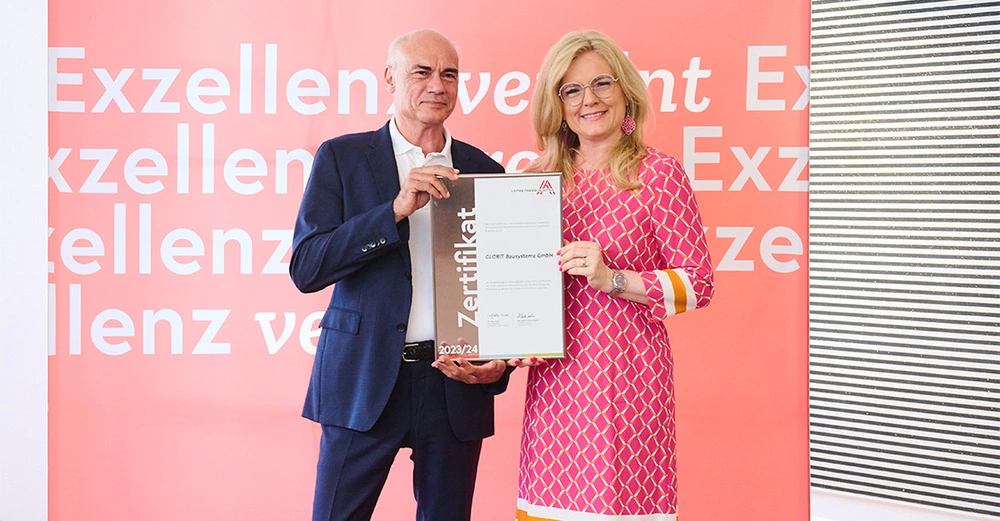 Zwei Personen halten ein Zertifikat vor einem Hintergrund mit dem Text Exzellenz. Das Zertifikat lautet Leittechnik Exzellenz Zertifikat GLOBRT Bussysteme GmbH 2023 24.