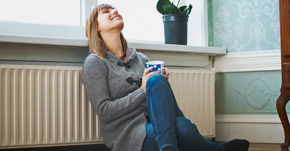 Eine Frau sitzt lächelnd am Boden und hält eine gepunktete Tasse in den Händen vor einer Heizung in einem gemütlichen Raum mit Tapeten und einem Fenster mit Topfpflanze.