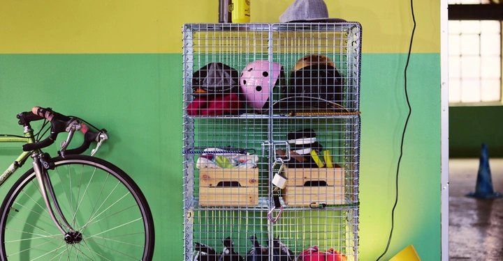 Ein silbernes Drahtregal mit Helmen und Schuhen steht in einem grünen Raum; daneben ein Fahrrad und eine gelbe Lampe auf dem Boden.