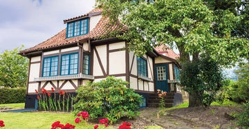 traditionelles Fachwerkhaus in weiß mit rotbraunem Fachwerk direkt an See. Wiese und Blumen im Vordergrund, perspektivische Aufnahme von links