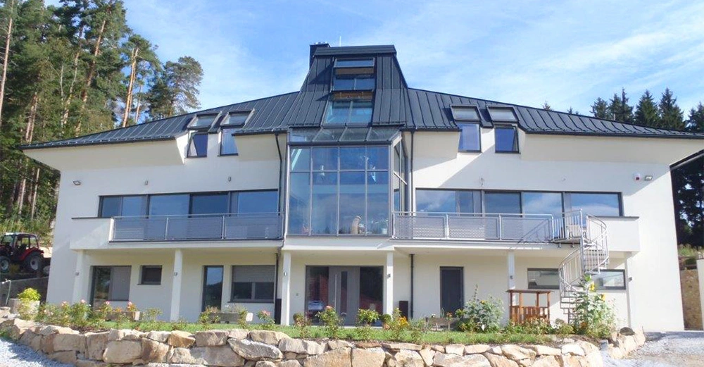 Großes modernes Gebäude steht still auf einem grasbewachsenen Gelände mit großen Glasfenstern, mehreren Balkonen und einem spiralförmigen Außentreppenhaus, von Wald umgeben.