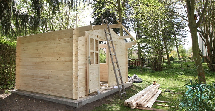 Holzhaus im Bau stehend auf Betonfundament mit angelehnter Leiter und umliegenden Brettern im grünen Garten mit Rasen und Bäumen bei Tageslicht