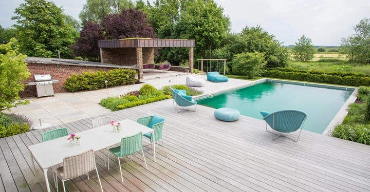 Terrasse mit Gartenmöbeln und Esstisch neben einem Swimmingpool in einem grünen, bewaldeten Garten. Grill und überdachter Loungebereich im Hintergrund, Landlandschaft in der Ferne sichtbar.