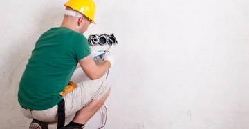 Ein Arbeiter kniet auf einem Pappstück und repariert elektrische Verkabelung an einer Wand im Innenbereich. Er trägt einen gelben Schutzhelm und Handschuhe, während verschiedene Werkzeuge neben ihm liegen.